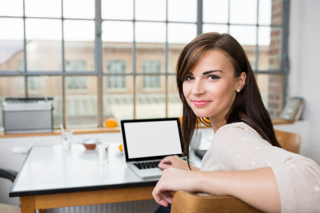 Attractive young woman with a cute smile turning in her chair to smile at the camera while sitting at a table with a laptop computer 